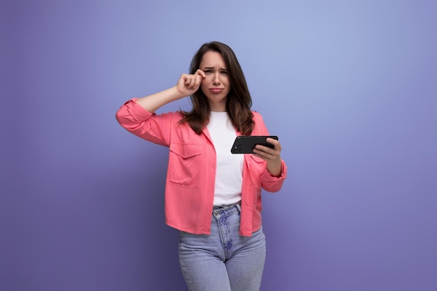 Thinking young woman in pink shirt and jeans with phone on studio isolated background