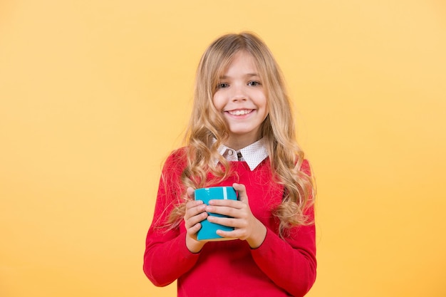 Thirst, dehydration concept. Health and healthy drink. Tea or coffee break. Girl with long blond hair in red sweater hold mug. Child smile with blue cup on orange background.