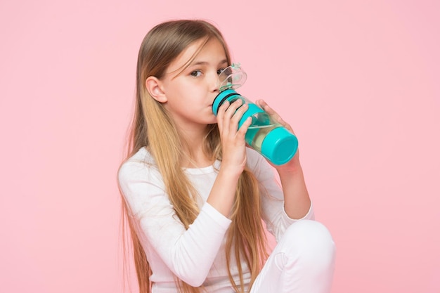 Фото Жаждущий ребенок пьет воду для здоровья на розовом фоне. детская бутылка с водой. маленькая девочка с пластиковой бутылкой. жажда и обезвоживание. детская активность и энергия, пробивной пастельный тренд.