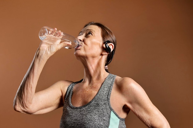 Thirsty senior woman in an active sportswear drinking water on a brown wall