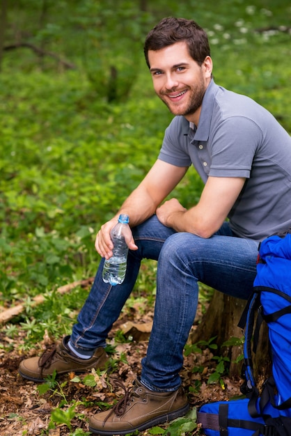 のどが渇いた旅行者。バックパックが彼の近くに横たわっている森の切り株に座っている間、水でボトルを保持しているハンサムな若い男
