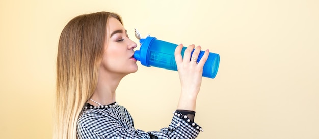 Thirsty woman with blue plastic bottle against a light background. A beautiful young caucasian businesswoman in a blouse drinking water on yellow.