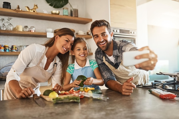 This calls for a selfie Shot of a happy young family posing for a selfie while cooking together in their kitchen