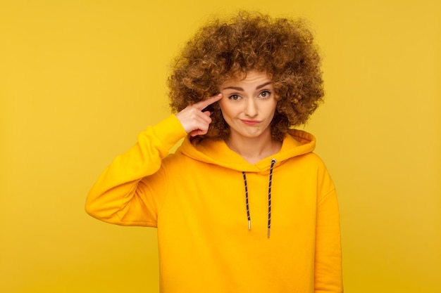 This is crazy. Portrait of curly-haired woman in urban style hoodie making stupid sign with finger near head, gesturing bad mind, dumb insane idea. indoor studio shot isolated on yellow background
