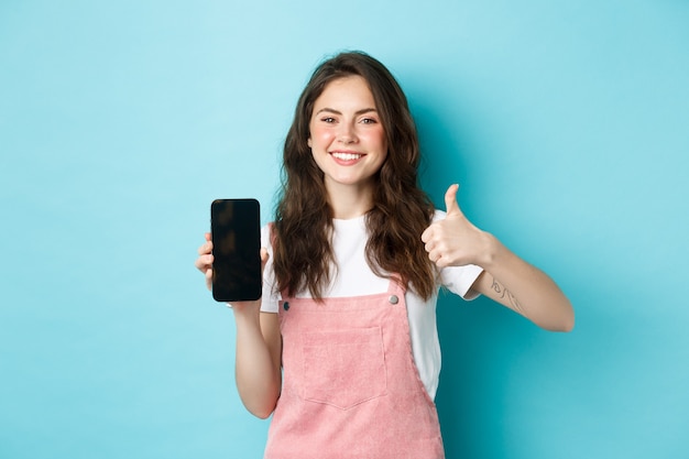 これはいい。笑顔の陽気な女性は、青い背景に立って、空のスマートフォンの画面と親指を上げてアプリやオンラインストアをお勧めします