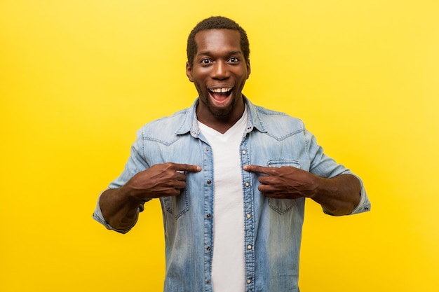 This is me. portrait of astonished young man in denim casual shirt pointing at himself, looking surprised by victory, rejoicing in achievement. indoor studio shot isolated on yellow background