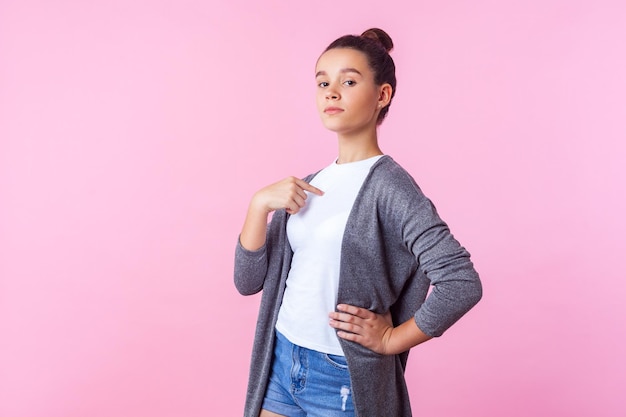 This is me Portrait of proud brunette girl with bun hairstyle in casual clothes pointing at herself looking arrogant and selfish egoistic teenager indoor studio shot isolated on pink background