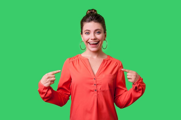 This is me. Portrait of successful woman with bun hairstyle, big earrings and in red blouse pointing at herself, looking proud and amazed about achievements. indoor, isolated on green background