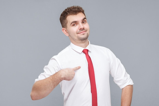 This is me. Portrait of young handsome proud man in white shirt and tie standing, pointing himself and looking at camera with proud face and confidence. indoor isolated on gray background.