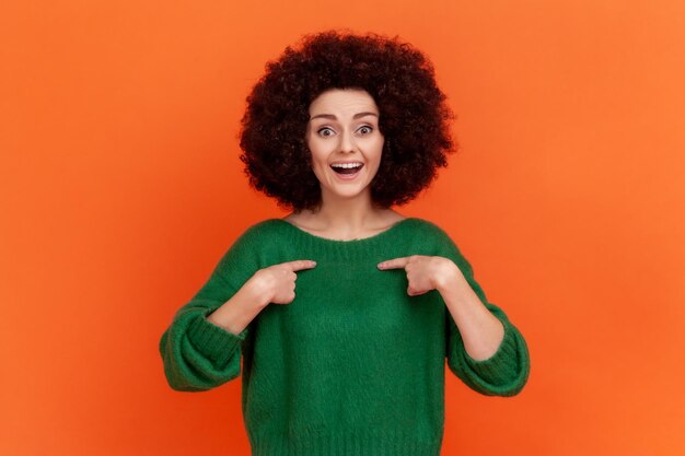 This is me Woman with Afro hairstyle wearing green casual style sweater with amazed face and pointing herself being proud and happy Indoor studio shot isolated on orange background