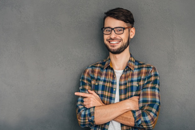 This is my choice! Cheerful young man in glasses pointing at copy space and looking at camera with smile while standing against grey background