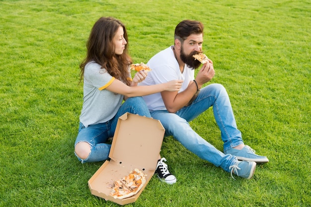 これは私のスライス夏のピクニックです緑の草でピザを食べる幸せなカップル家族の週末のカップルの恋にファーストフードのひげを生やした男のヒップスターと愛らしい女の子がピザを食べる