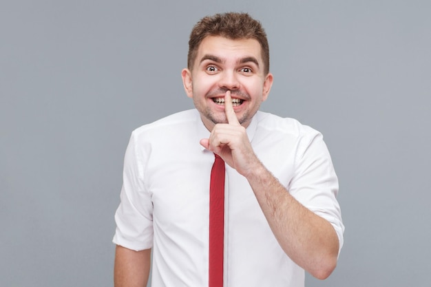 This is secret Portrait of young handsome man in white shirt and tie standing with finger on lips tell secret and smiling indoor isolated on gray background