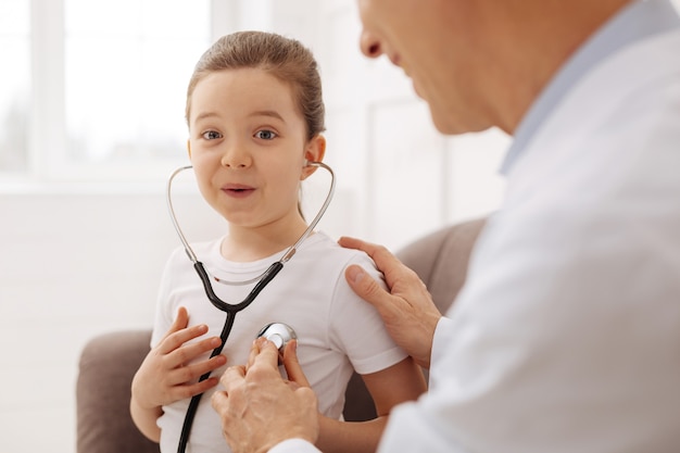 This is so cool. Adorable curious young child having fun with doctor using his stethoscope for listening to her own heart