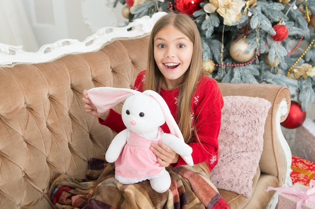 This is unbelievable. Little girl with cute bunny at Christmas tree. Small child happy smiling with present. Small girl hold rabbit toy. Little child play with soft toy. Best Christmas toy.