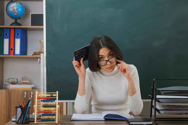 thiunking kijken naar camera jonge vrouwelijke leraar die een bril draagt en een rekenmachine vasthoudt die aan een bureau zit met schoolhulpmiddelen aan in de klas
