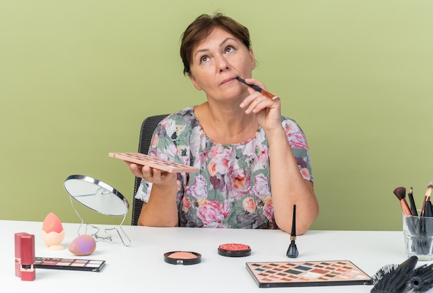 Thoughtful adult caucasian woman sitting at table with makeup tools holding eyeshadow palette and makeup brush looking up isolated on olive green wall with copy space