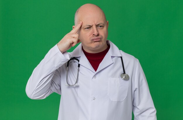 Foto uomo slavo adulto premuroso in uniforme da medico con stetoscopio che mette il dito sulla tempia e guarda di lato