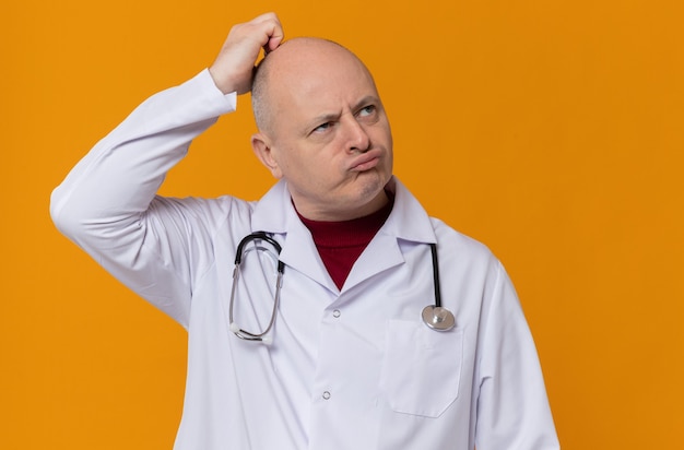 Thoughtful adult slavic man in doctor uniform with stethoscope scratching his head and looking up