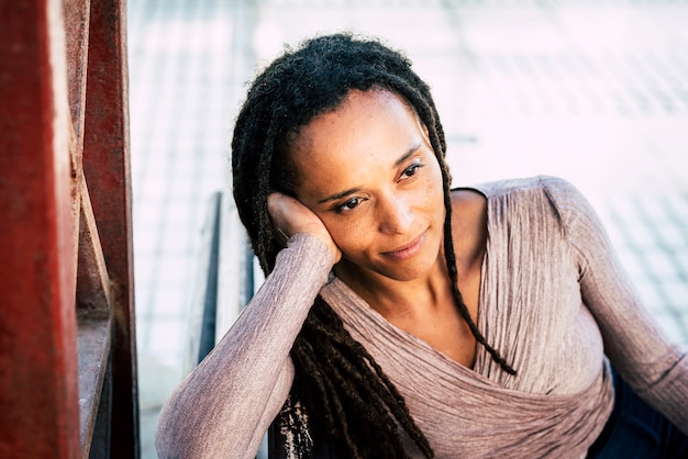 Thoughtful african american woman smiling with head in hand. Black woman with deadlocks hairstyle. Young woman sitting on park bench. Stylish woman with dreadlocks daydreaming outdoors on bench