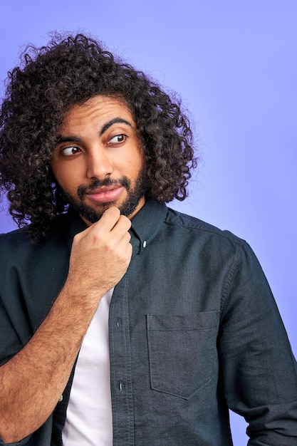 Thoughtful arabic male with curly long hair standing in contemplation and looking away, holding hand on chin, isolated over purple wall