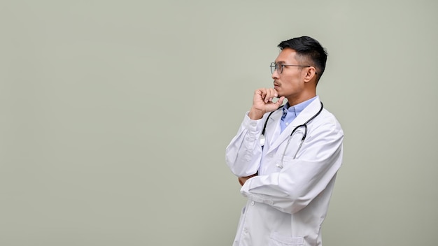 Thoughtful Asian male doctor in a white gown and glasses hand on chin looking aside