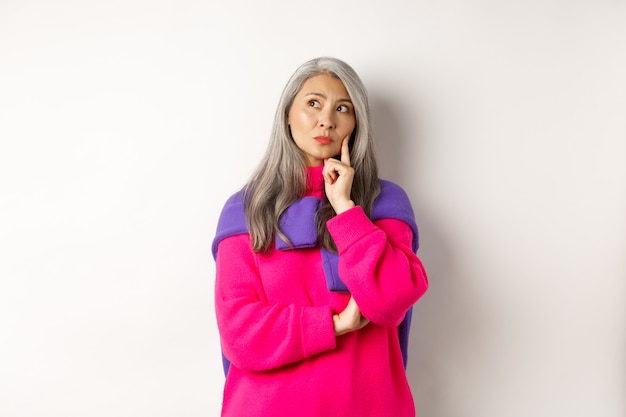 Photo thoughtful asian middle-aged mother looking upper left corner, thinking with serious face, standing over white background. copy space