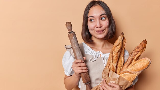 Thoughtful asian woman baker holds paper bag of fresh crunchy\
baguettes and rolling pin smeared with flour wears apron isolated\
over beige background blank space for your promotional content