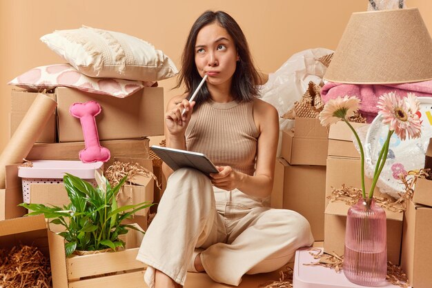 Thoughtful asian woman prepares designing project on tablet\
draws sketches with stylus imagines her new apartment for living\
wears t shirt and trousers collects personal belongings in carton\
boxes