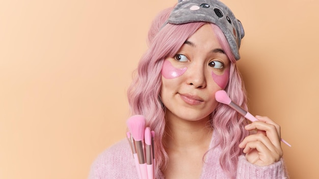 분홍색 머리를 염색한 사려 깊은 아시아 여성은 화장용 브러시를 사용하여 얼굴 파우더를 바르고 눈 밑에 하이드로겔 패치를 바르고 갈색 벽에 집중된 수면 마스크를 착용합니다