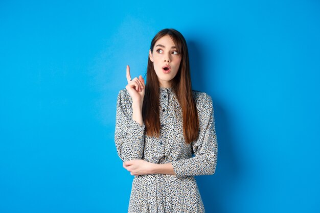 Thoughtful attractive girl in dress pitching an idea, raising finger and looking up, have a plan, standing on blue.