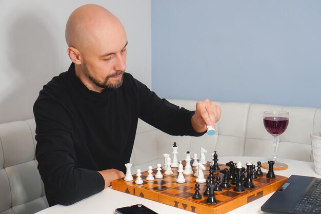 Thoughtful bald man wearing beard playing chess on board near\
laptop and wineglass