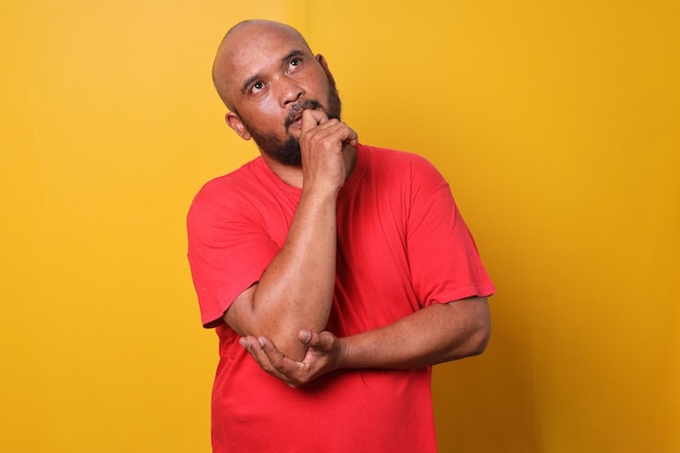 Thoughtful bearded bald man in red tshirt looking aside and thingking over yellow background