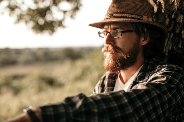 Thoughtful bearded male in eyeglasses sitting in green field