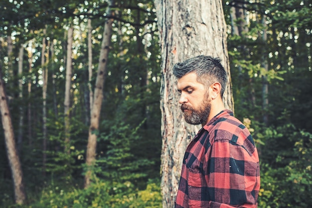 Thoughtful bearded man in lumberjack shirt alone in forest or park Handsome hiker enjoying nature on his own Active lifestyle traveling and solitude concepts