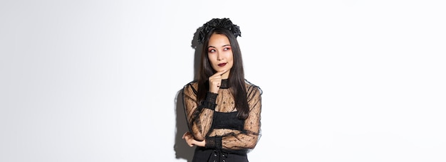 Thoughtful beautiful asian woman standing in black lace dress and gothic wreath looking upper left c