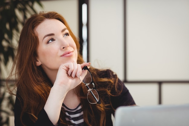 Thoughtful beautiful woman holding eyeglasses while looking up at creative office