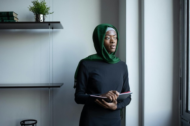 ヒジャブを着た考え深い黒人イスラム教徒の女性がノートブックを持って立っています