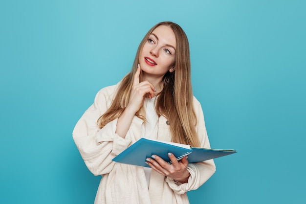 Thoughtful blonde student girl holding an open exercise book and looking to copy space isolated on blue wall 