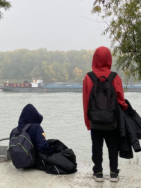 Thoughtful boys with hoods looking a cago ship sailing down Danube river Cold and calm in winter day