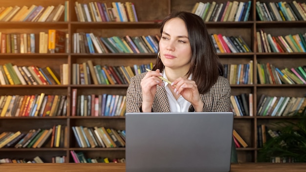 Thoughtful brunette in brown jacket over white blouse holds pen sitting at grey laptop against coloured books on racks and pot plant in library, sunlight