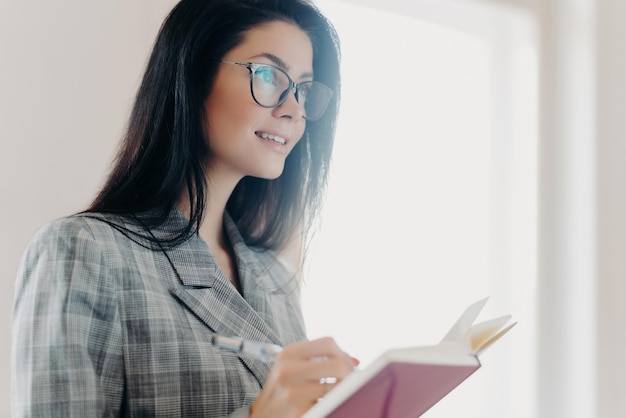 Thoughtful brunette European woman in formal attire and transparent glasses writes down text information in notepad deep in concentration indoors