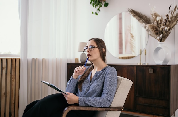 a thoughtful brunette psychologist woman with glasses is sitting in a chair with papers in the room and taking notes