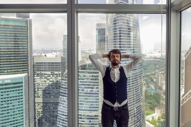 수염을 기른 사려 깊은 사업가는 흰 셔츠와 조끼를 입은 모스크바의 고층 빌딩을 배경으로 사무실 창문 옆에 서 있다