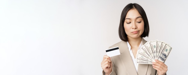 Thoughtful businesswoman korean corporate woman showing credit card and money cash dollars in hands standing over white background and thinking