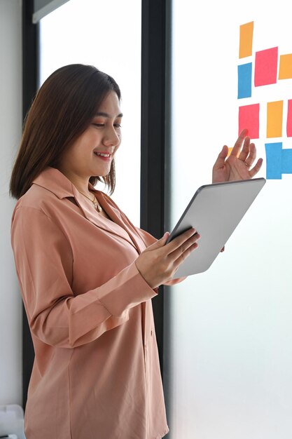 Thoughtful businesswoman reading sticky notes on the glass wall in office.