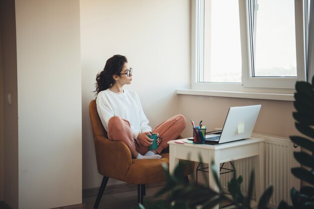 窓の近くのPCの前でお茶を飲む巻き毛と眼鏡を持つ思いやりのある白人ビジネスの女の子