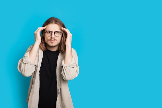 Thoughtful caucasian man with long hair and eyeglasses is touching his head and looking at camera on a blue studio wall with free space