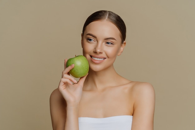 Фото Задумчивая очаровательная европейка держит яблоко возле лица, нежно улыбается, имеет белые идеальные зубы, здоровая чистая кожа, завернутая в подставки для полотенец для душа с обнаженными плечами