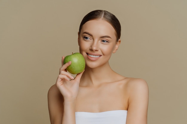 La donna europea affascinante e premurosa tiene la mela vicino al viso sorride delicatamente ha denti bianchi perfetti e una pelle pulita